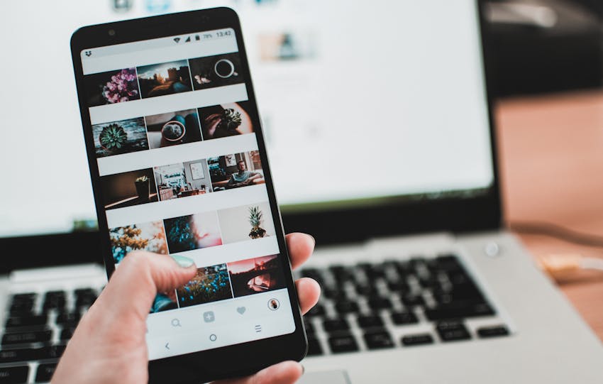 A person uses a smartphone to browse social media in front of a laptop, emphasizing modern technology and lifestyle.