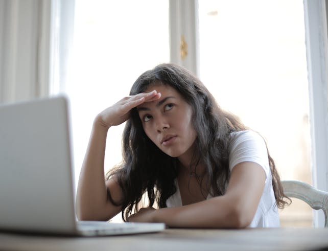 Irritated ethnic female entrepreneur in casual wear sitting at table with netbook and touching head while waiting for internet connection during remote work